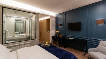 A modern hotel room with a large bed, a desk with a chair, a TV mounted on the wall, and a bathroom with a large mirror and a sink.