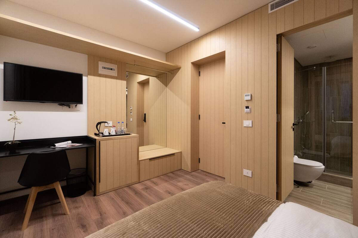 A hotel room with a bed, a desk with a chair, a television, a shower, and a toilet.