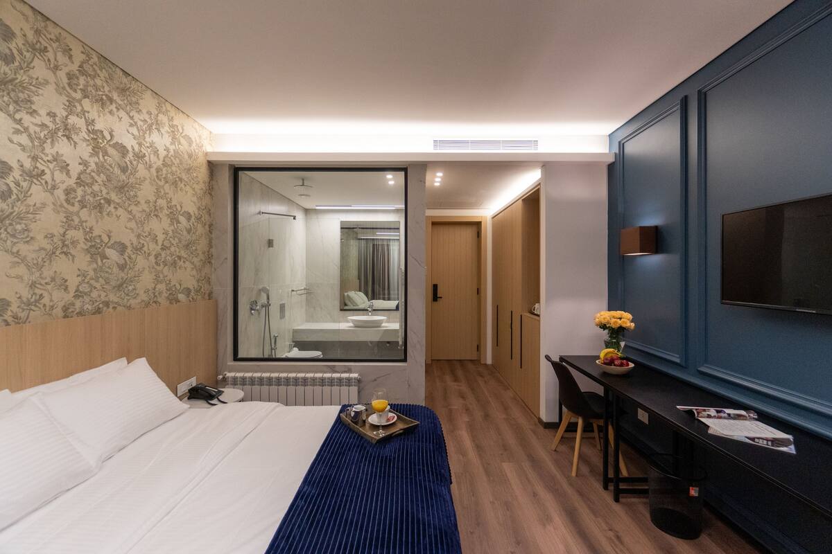 A modern hotel room with a large bed, a desk with a chair, a TV mounted on the wall, and a bathroom visible through a glass door.