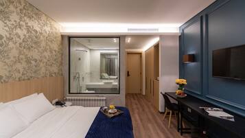 A modern hotel room with a large bed, a desk with a chair, a TV mounted on the wall, and a bathroom visible through a glass door.