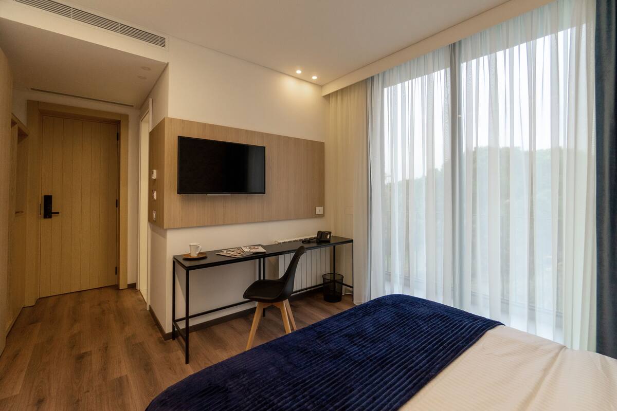 A modern hotel room with a bed, a desk, a TV, and a large window with curtains.