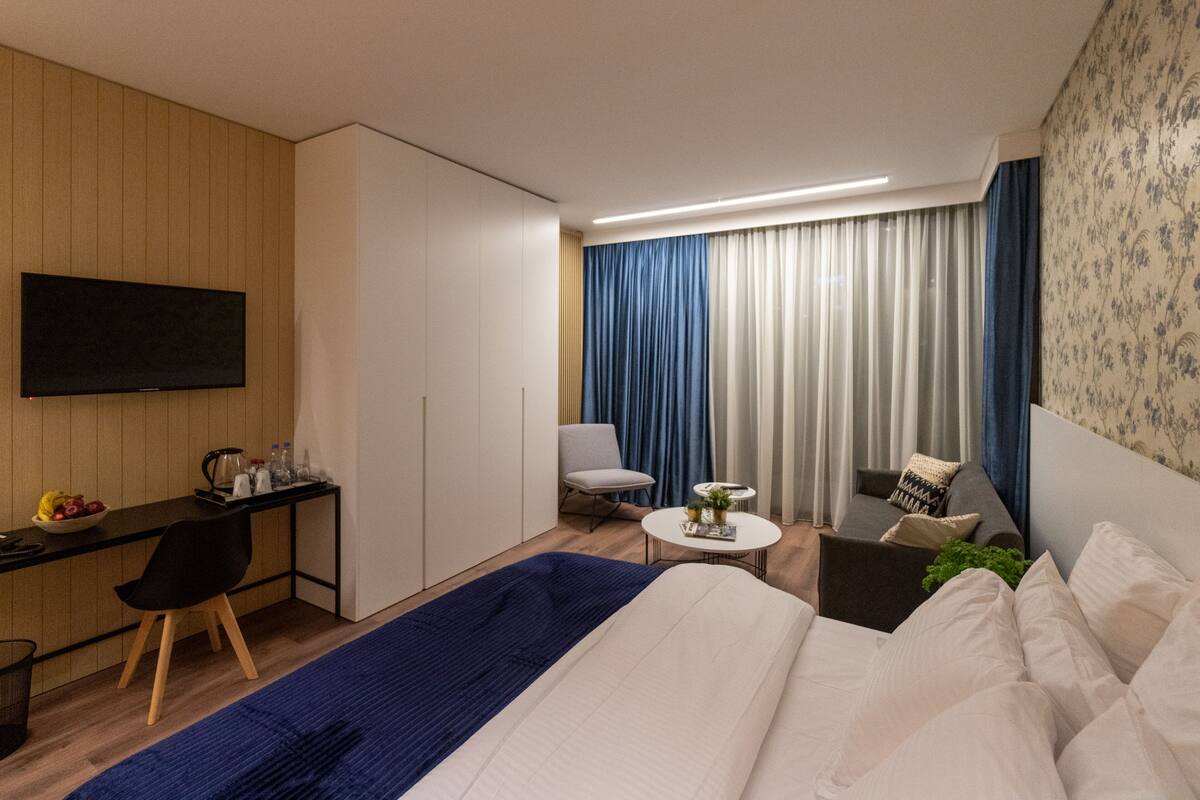 A modern hotel room with a bed, a desk, a chair, a small table, and a TV.