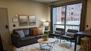 A modern living room with a sofa, chairs, a coffee table, and a large window with a city view. - Modern Luxe Condo | Walk to Downtown TC | State 210 (Traverse City)