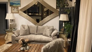 Una sala de estar con un sofá gris, una mesa de centro de madera con un florero, un espejo grande y un televisor sobre un mueble.