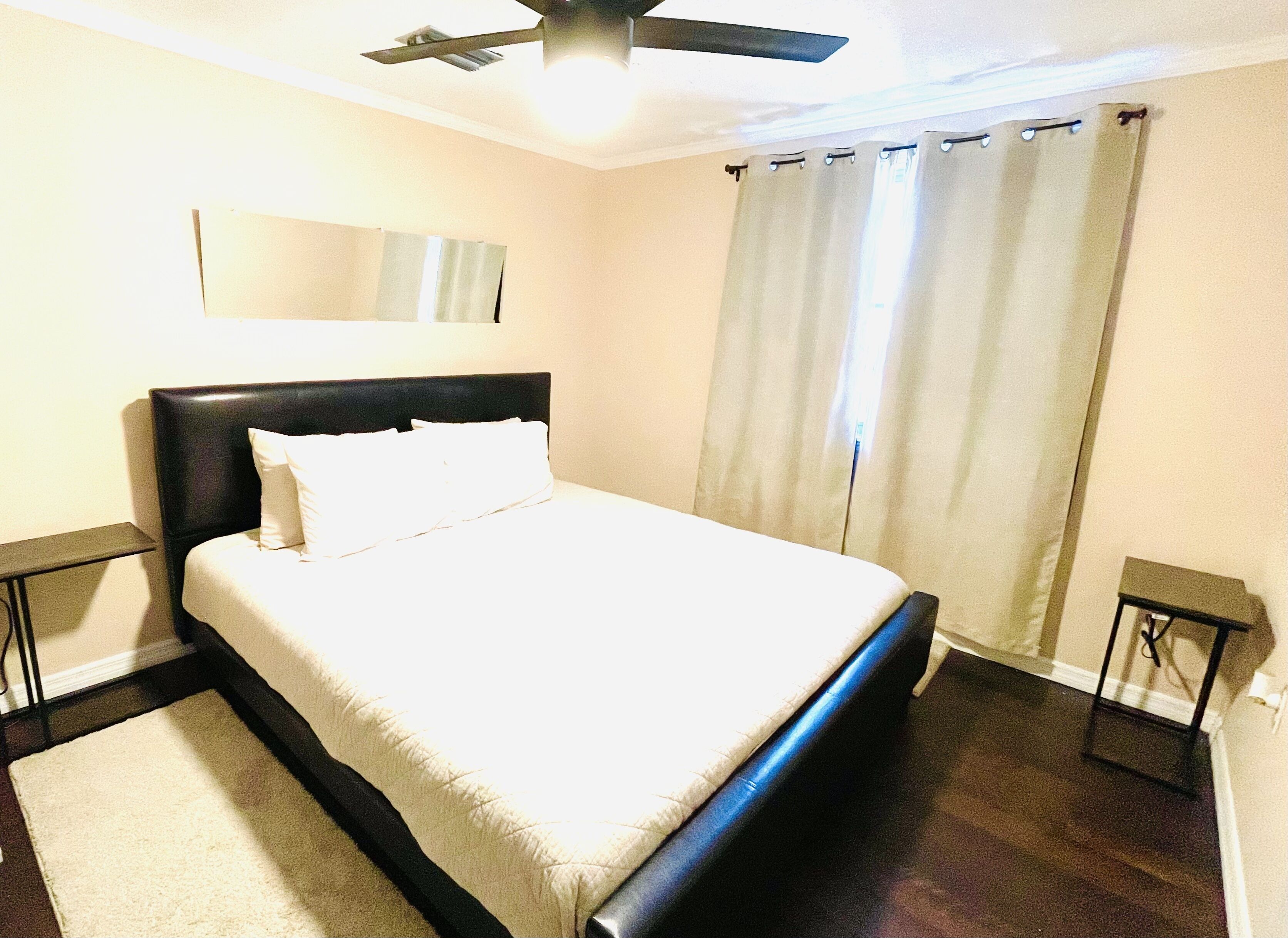 Un letto rifatto con lenzuola bianche in una stanza con ventilatore a soffitto, un'ampia finestra con tende e un comodino accanto al letto.
