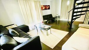 Ein Wohnzimmer mit einer schwarzen Couch, einer weißen Couch, einem Glastisch und einem kleinen Tisch mit Blumen in einer Vase. Eine Treppe befindet sich rechts und ein Gemälde ist an der Wand zu sehen.