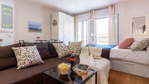 Ein Wohnzimmer mit Sofa, Couchtisch und einem Bett mit Kissen. Es gibt ein Fenster mit Vorhängen, ein Gemälde an der Wand und eine Uhr.