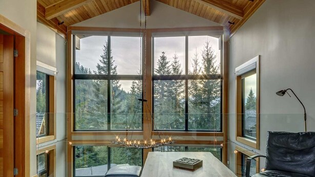 Une pièce dotée d’une grande fenêtre donnant sur des arbres et un ciel nuageux. On y trouve une table en bois avec un coussin, une chaise en cuir noir et un luminaire suspendu.