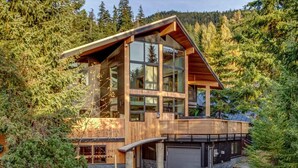 Une maison moderne en bois, dotée de grandes fenêtres, d’un garage et d’une terrasse, entourée d’une forêt dense.