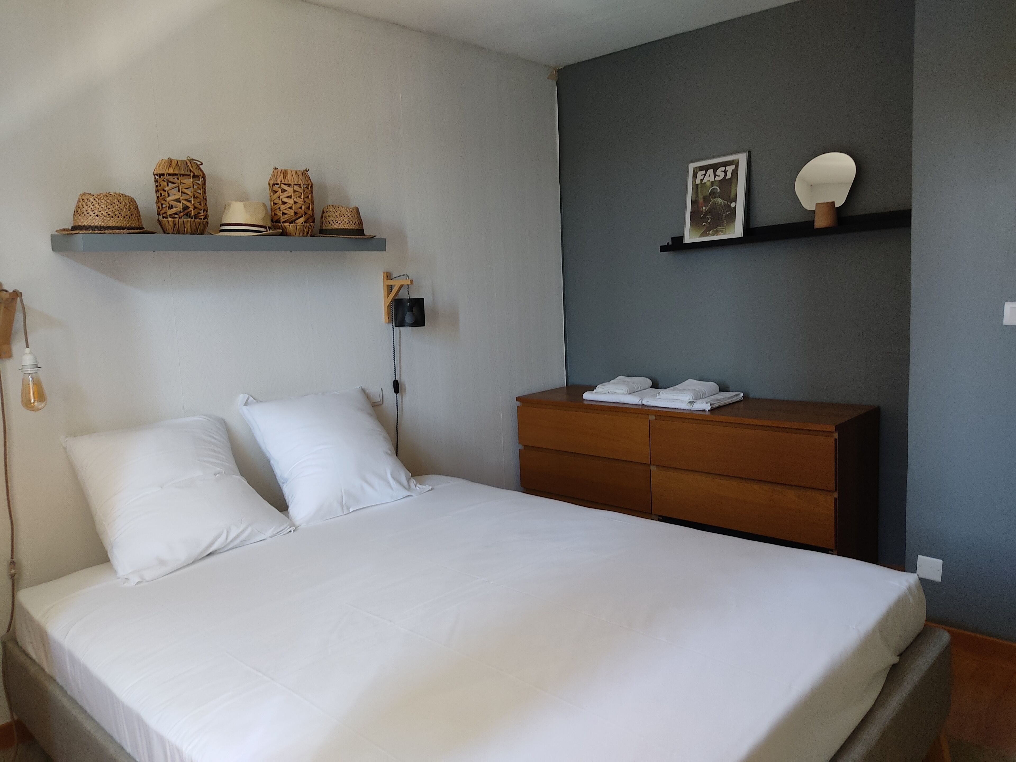 A neatly made bed with white linens, a wooden dresser with folded towels, and decorative baskets on a shelf above the bed.