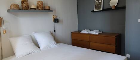 A neatly made bed with white linens, a wooden dresser with folded towels, and decorative baskets on a shelf above the bed.