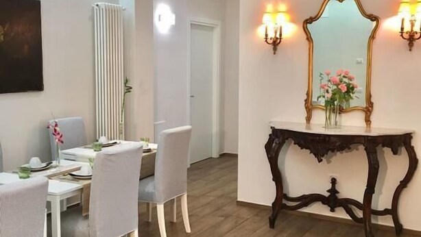 Een eetgedeelte met een witte tafel en stoelen, een houten console met een spiegel en wandlampen, en een radiator aan de muur.