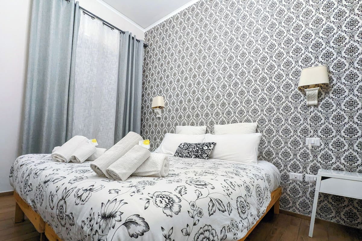 Un lit bien fait, avec une literie à motifs floraux, une table de chevet et un mur recouvert d’un papier peint à motifs.