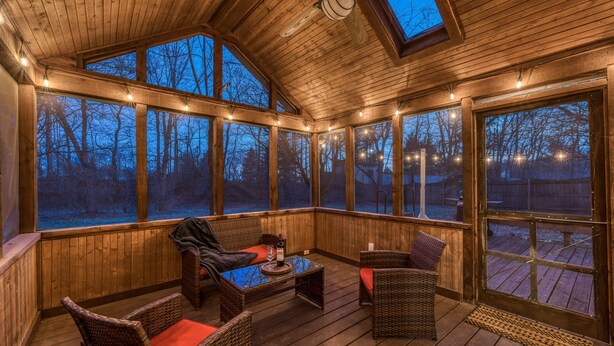 A covered outdoor area with wooden flooring, wicker furniture, and large windows offering views of a wooded area. - Downstairs BR! Pet Friendly house w/4BR, fenced yard & Sun Room (Indianapolis)