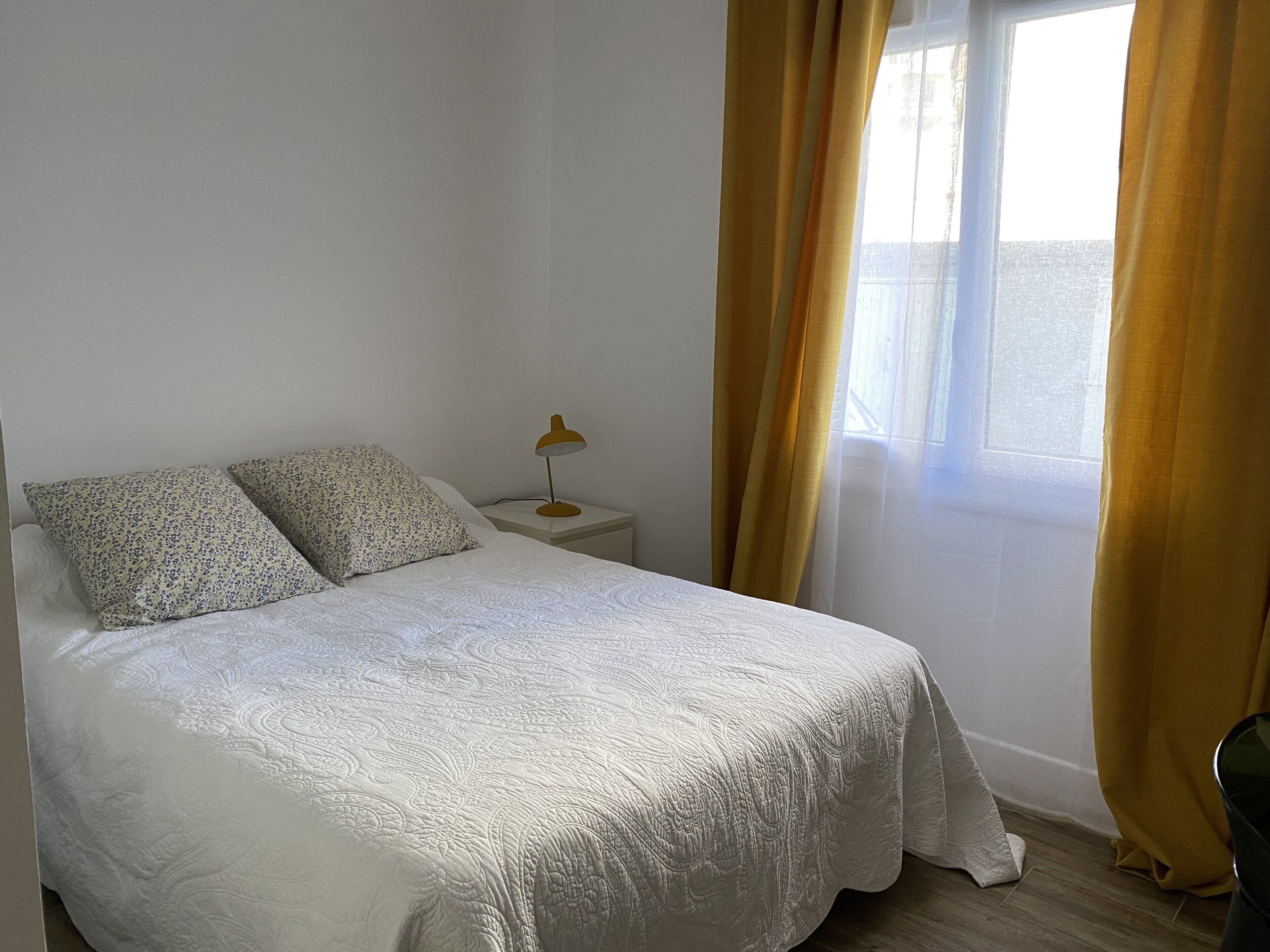 Un lit bien fait, recouvert d’une couvre-lit à motifs, avec deux oreillers, une lampe de chevet et une fenêtre ornée de rideaux jaunes.