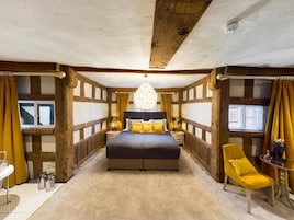 Quarto com cama grande, lustre, vigas de madeira e uma cadeira amarela.