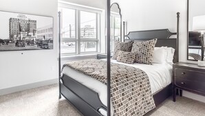 A four-poster bed with a patterned bedspread, a nightstand with a lamp, and a large window with a cityscape view.