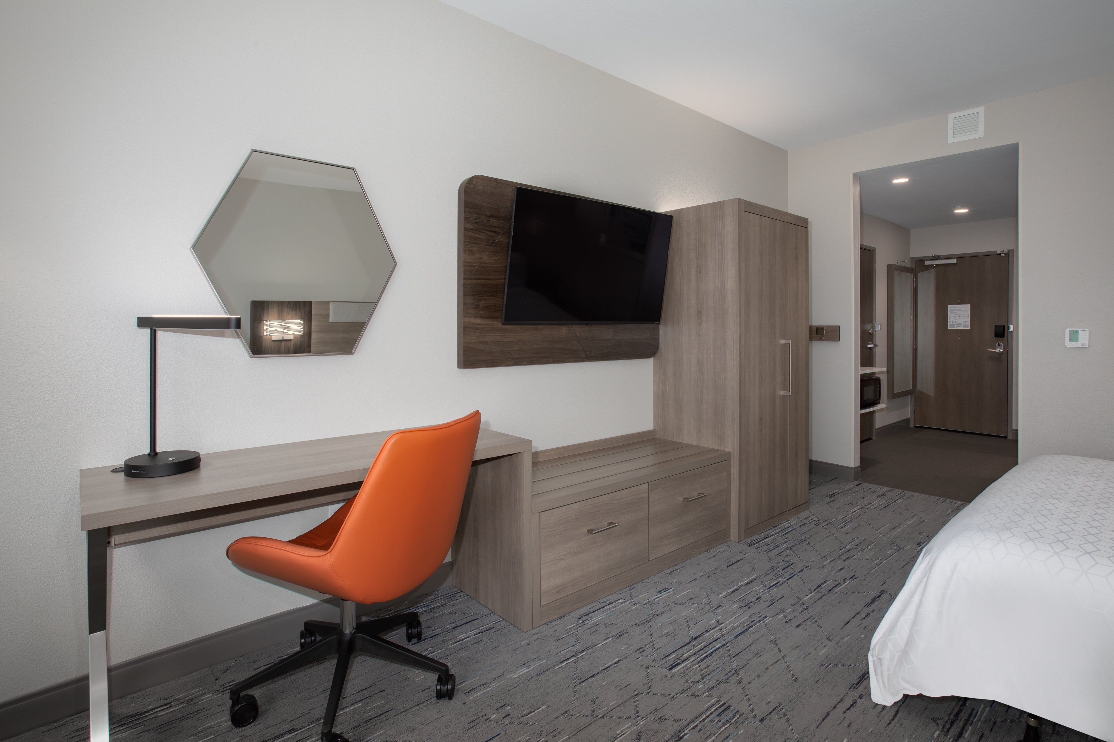 Ein modernes Hotelzimmer mit Schreibtisch, einem orangefarbenen Sessel, einem Bett, einem an der Wand montierten Fernseher und einem Kleiderschrank.