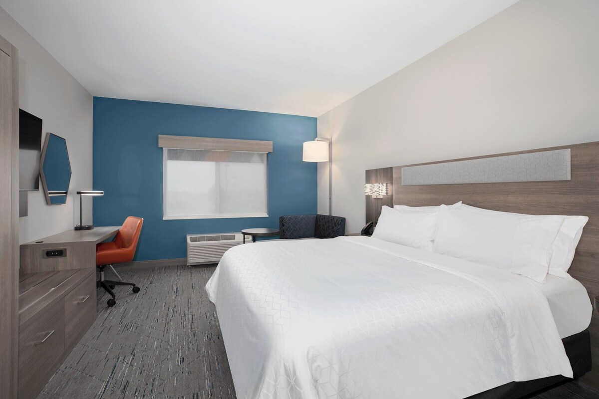 A hotel room with a large bed, a desk with a chair, a window with blinds, a wall-mounted lamp, and a radiator.