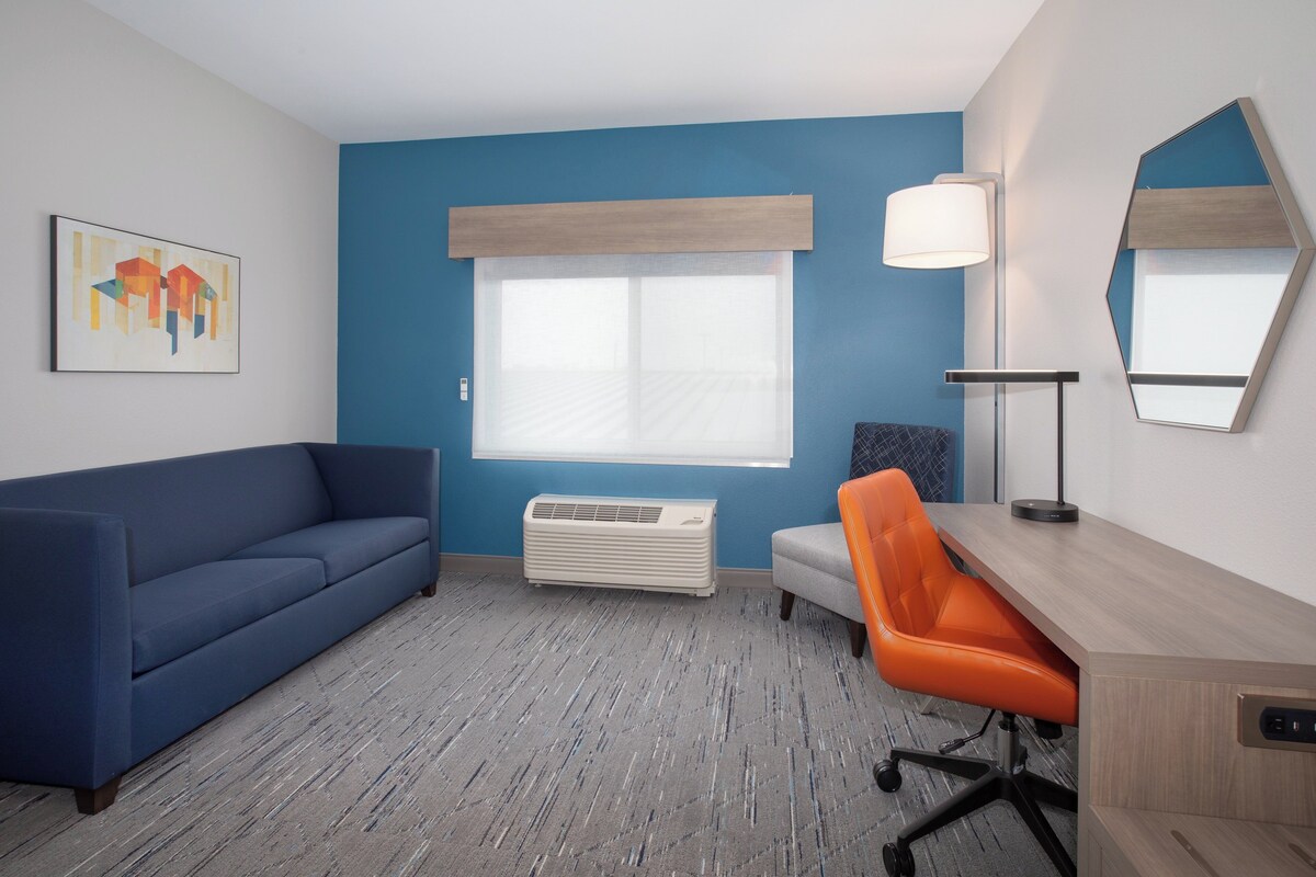 A room with a blue wall, a blue sofa, an orange chair, a desk, a lamp, and a large window with blinds.