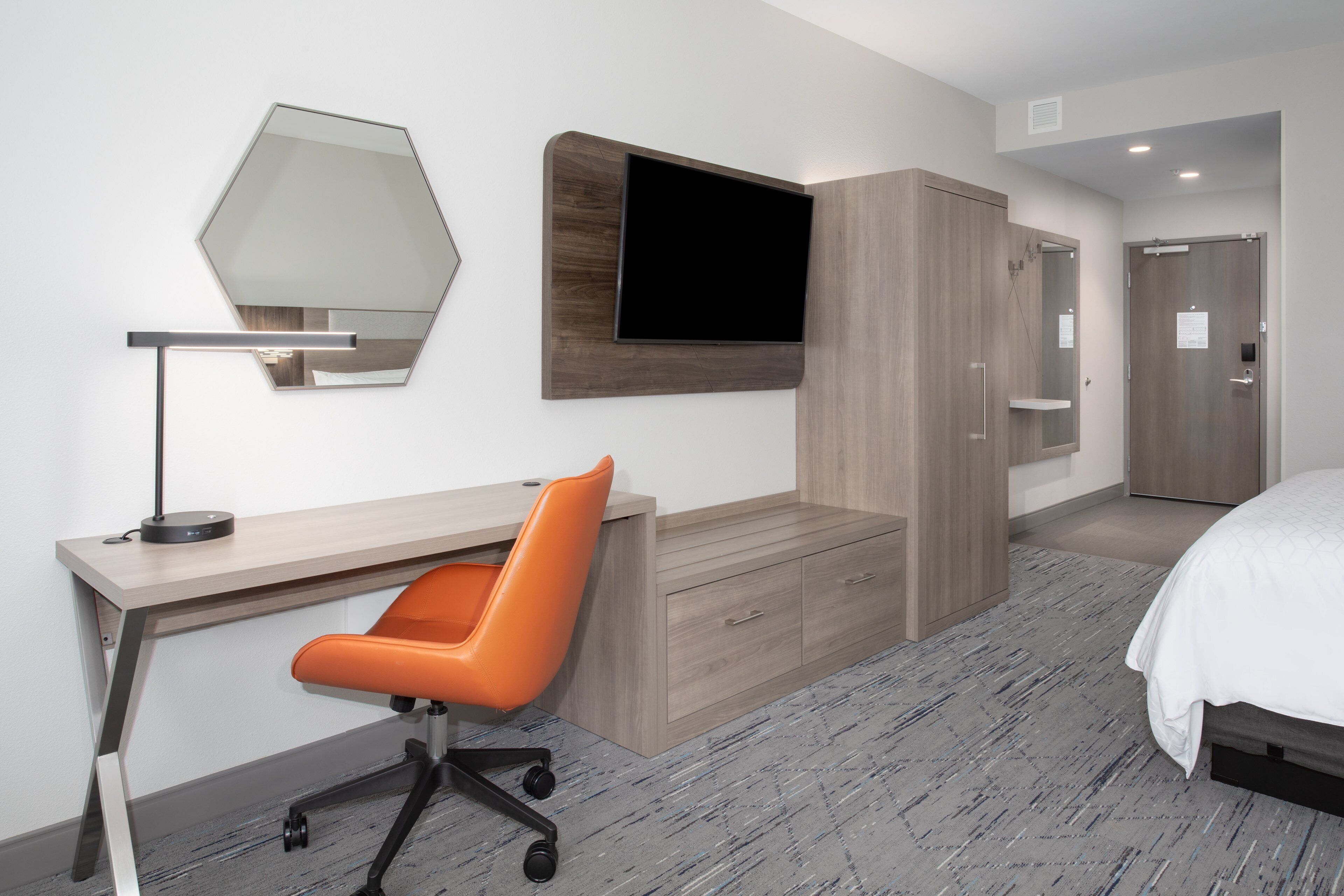 A modern hotel room with a wooden desk, an orange office chair, a flat-screen TV mounted on the wall, a wardrobe, and a bed with a white duvet.