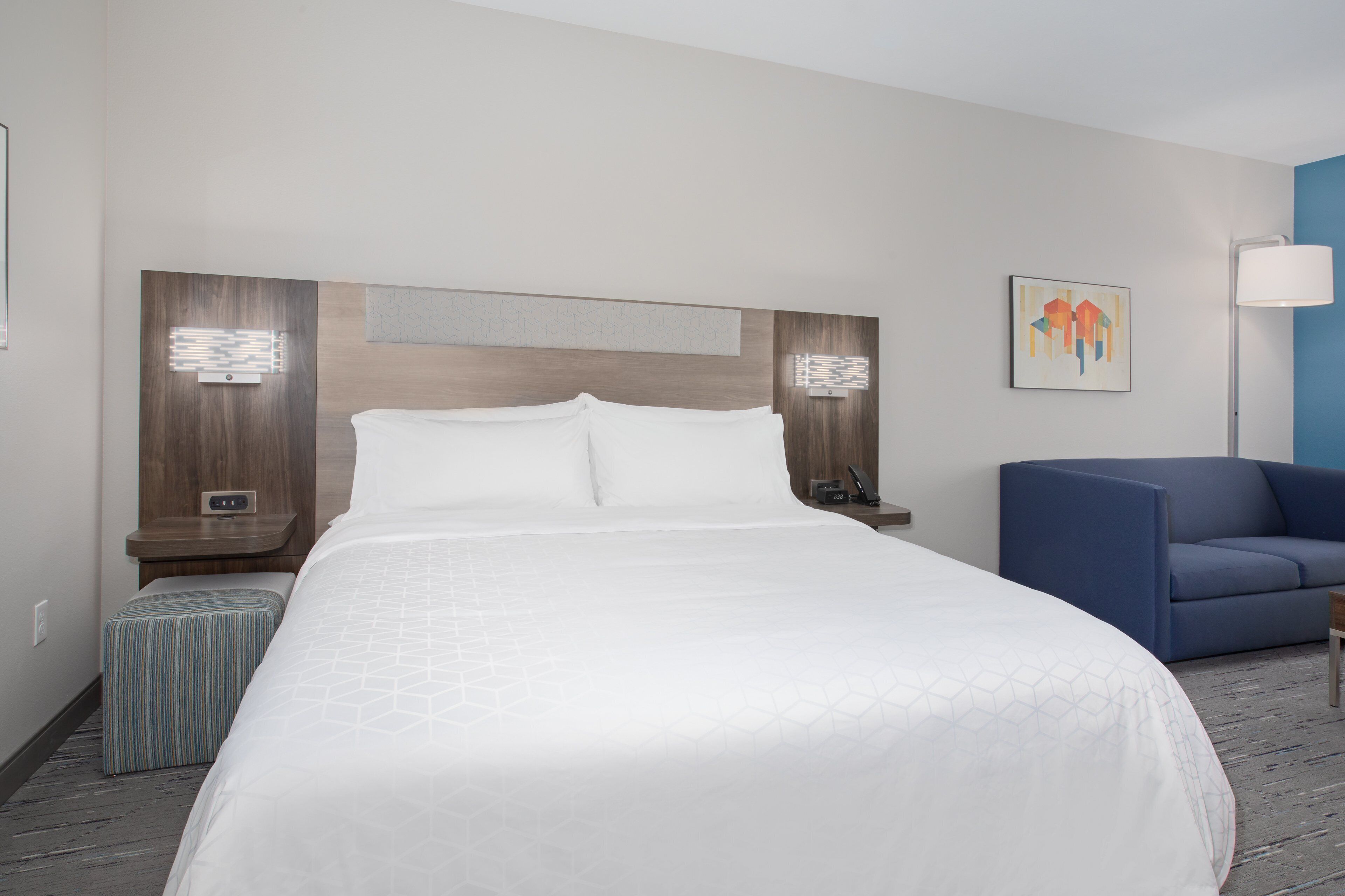 A hotel room with a large bed, a bedside table, a lamp, a chair, and a wall-mounted artwork.