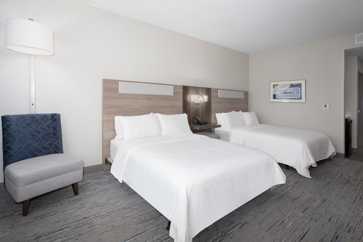 A hotel room with two beds, a small seating area, a lamp, and a wall-mounted artwork.