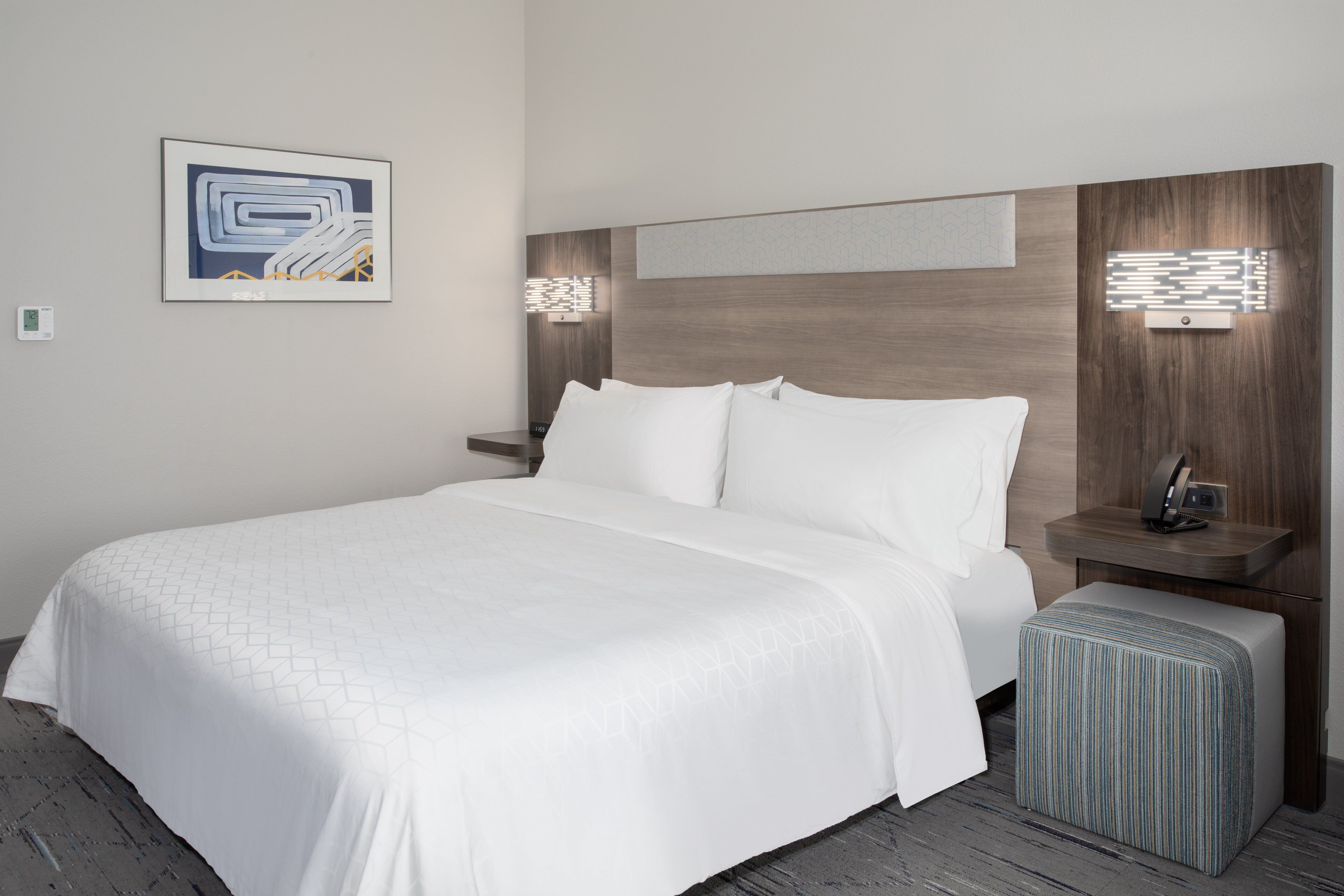 A modern hotel room with a large bed, a nightstand, a bedside table, and a wall-mounted light fixture.