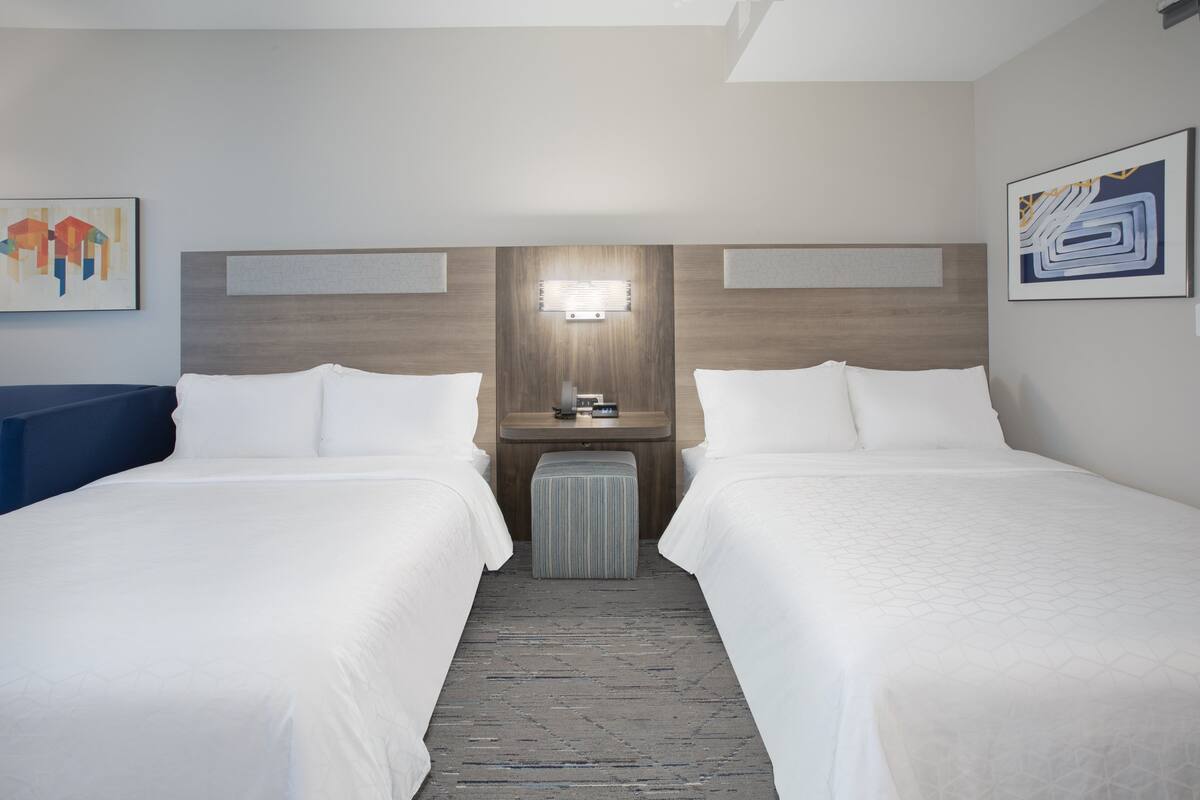 Hotellihuone, jossa on kaksi sänkyä, pieni pöytä lampun kanssa ja taideteoksia seinillä.