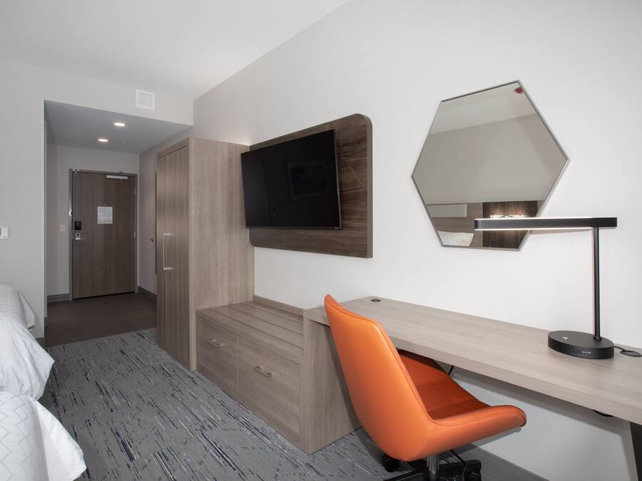 A modern hotel room with a bed, a desk with a chair, a flat-screen TV, and a large mirror.
