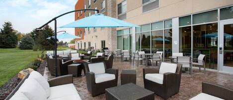 An outdoor patio area with wicker furniture, a blue umbrella, and a building with large windows in the background.