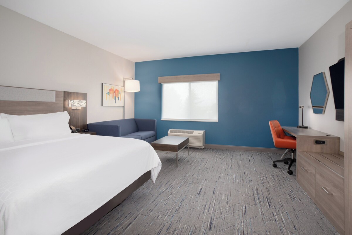 A hotel room with a large bed, a desk, a chair, a sofa, a window, and a wall-mounted TV.