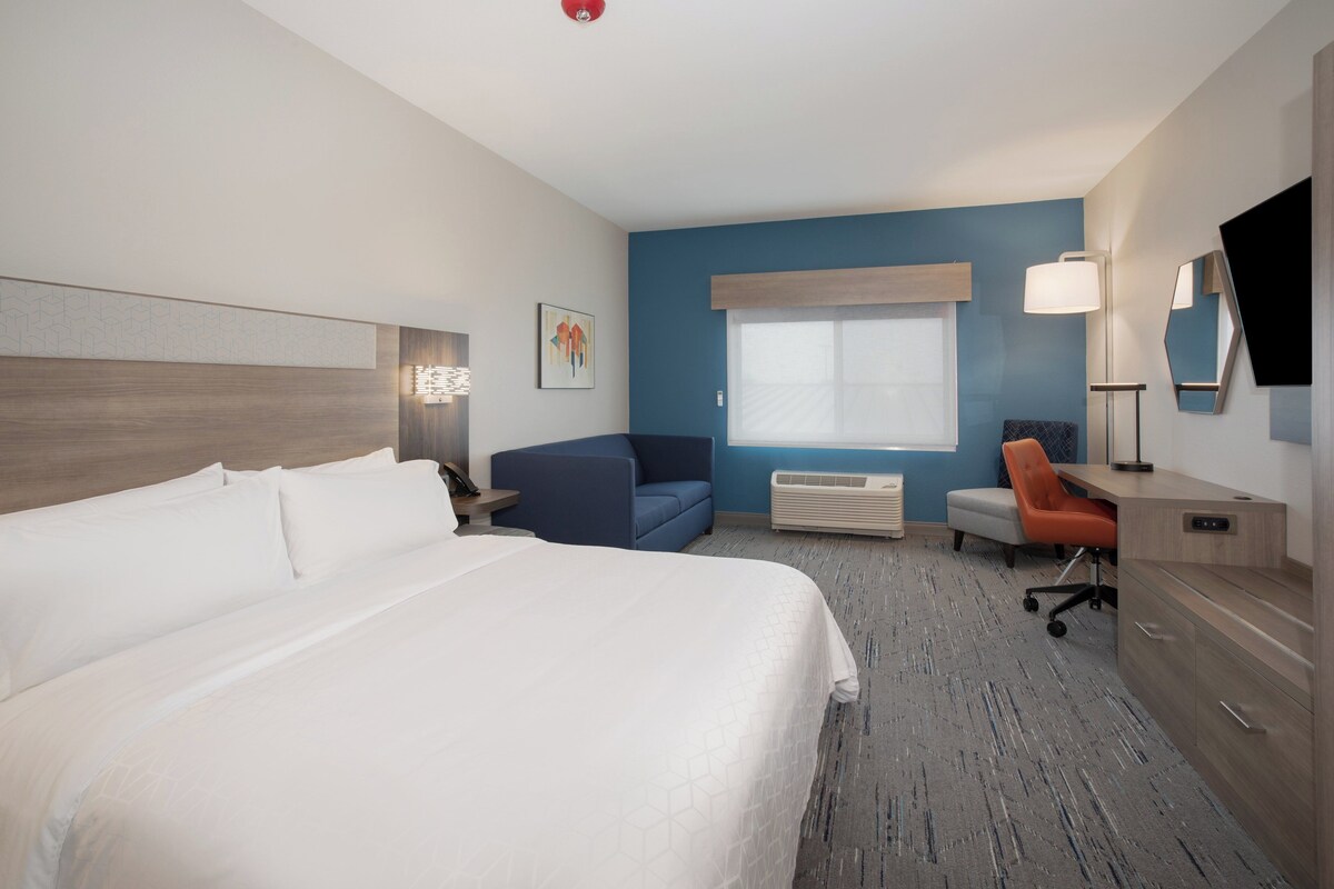 A hotel room with a large bed, a desk, a chair, a desk lamp, and a window with blinds.
