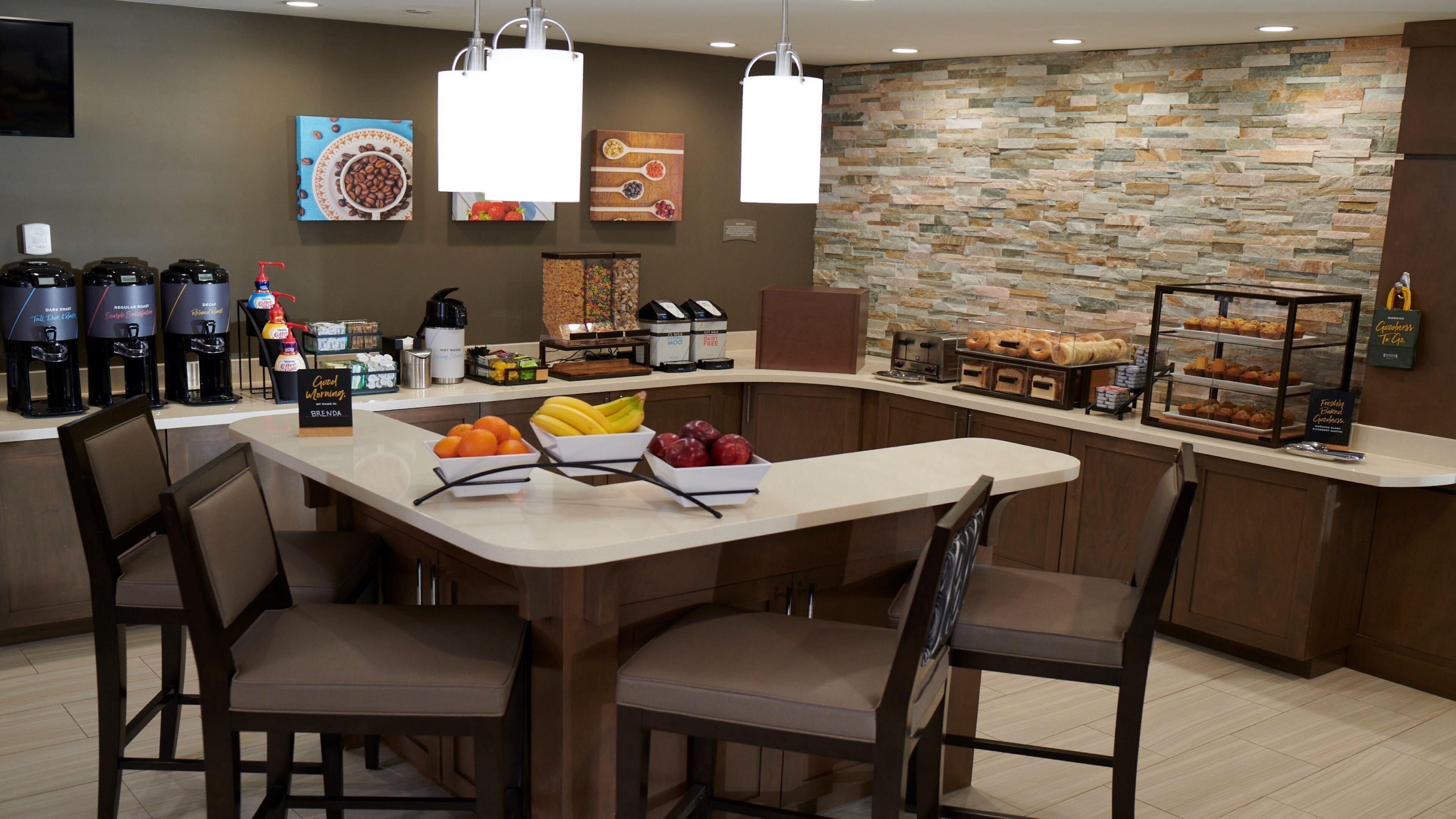 A breakfast bar with coffee makers, fruit bowls, and a stone wall.