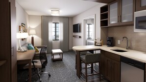 A compact hotel room with a kitchenette, a desk with a chair, a small table, a sofa, a TV mounted above the TV, and a window with curtains. - Staybridge Suites Southgate Detroit Area by IHG (Southgate)
