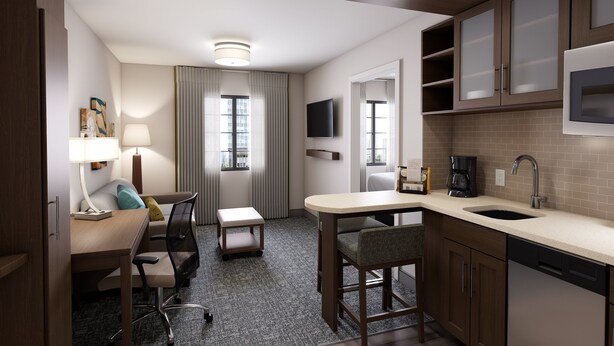 A compact hotel room with a kitchenette, a desk with a chair, a small table, a sofa, a TV mounted above the TV, and a window with curtains. - Staybridge Suites Southgate Detroit Area by IHG (Southgate)