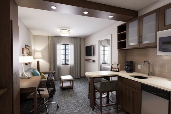 A compact hotel room with a kitchenette, a desk with a chair, a small table, a sofa, a TV mounted above the TV, and a window with curtains. - Staybridge Suites Southgate Detroit Area by IHG (Southgate)