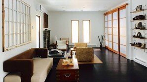 Uma sala de estar espaçosa com um sofá, uma mesa de centro e uma poltrona. Há duas janelas com persianas verticais, uma lareira e um sapateiro encostado na parede.