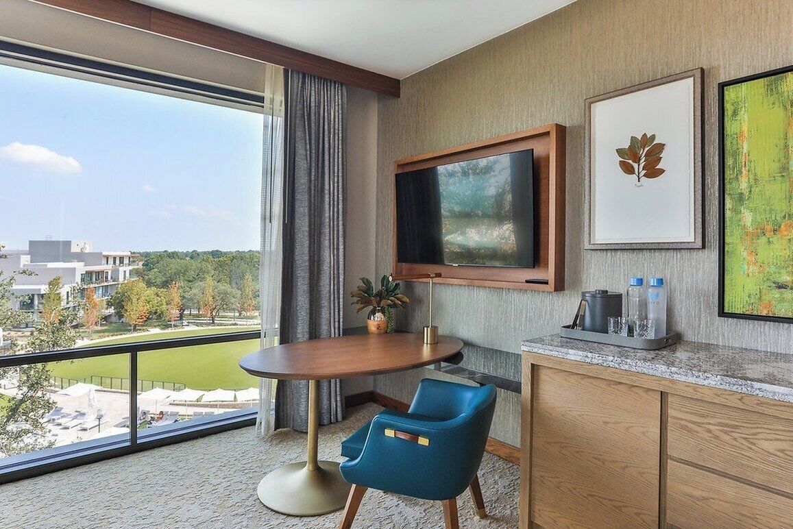 Een hotelkamer met een groot raam, een aan de muur bevestigde televisie, een tafeltje, een blauwe stoel en een houten kast.