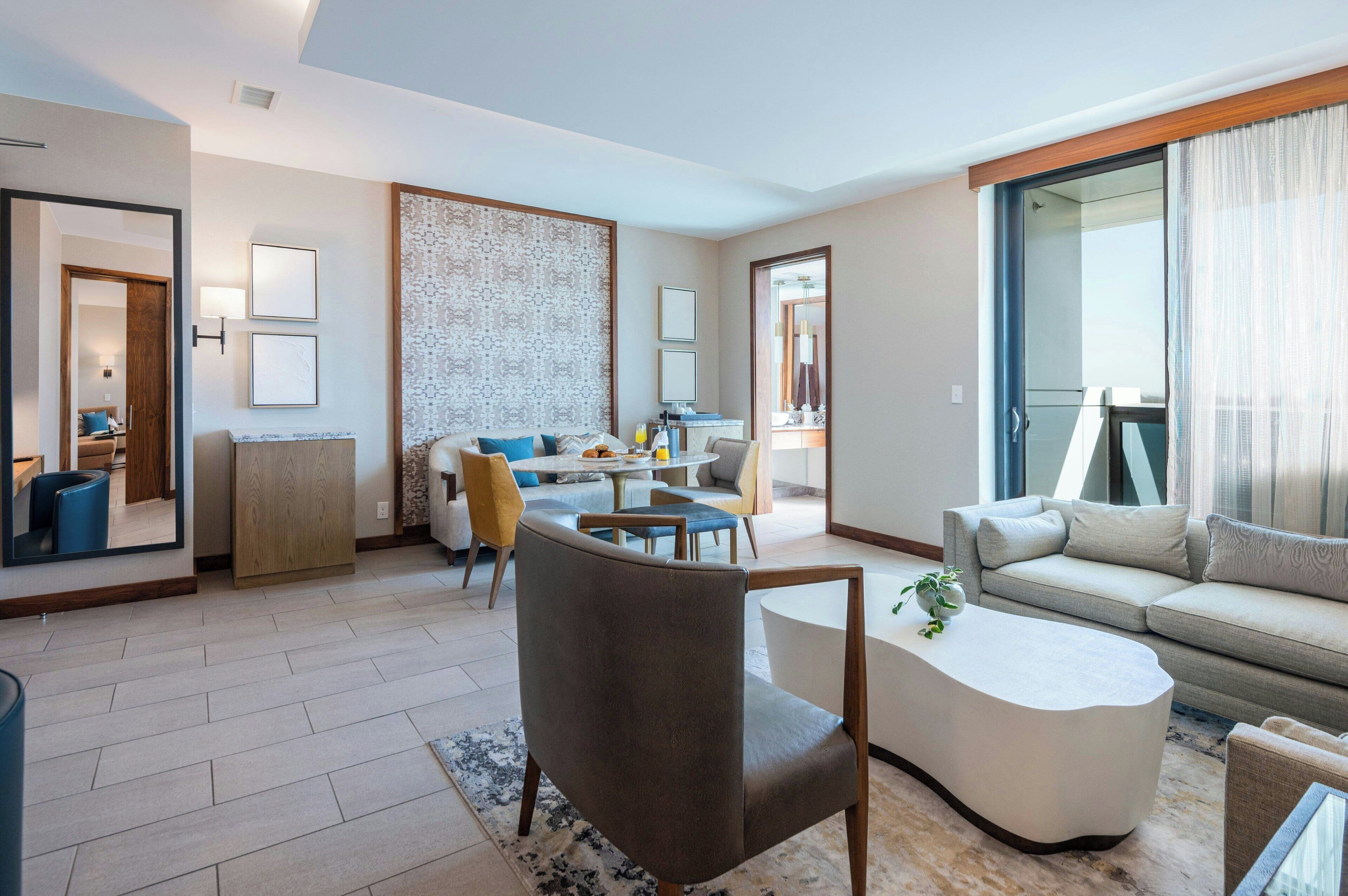 A modern hotel room with a living area, dining space, and a balcony with curtains.