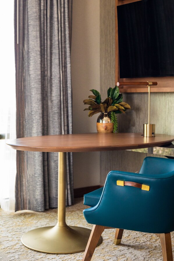 Una mesa redonda de madera con base dorada, una silla verde aguamarina con correas de cuero, una planta en maceta y una pantalla negra en la pared.