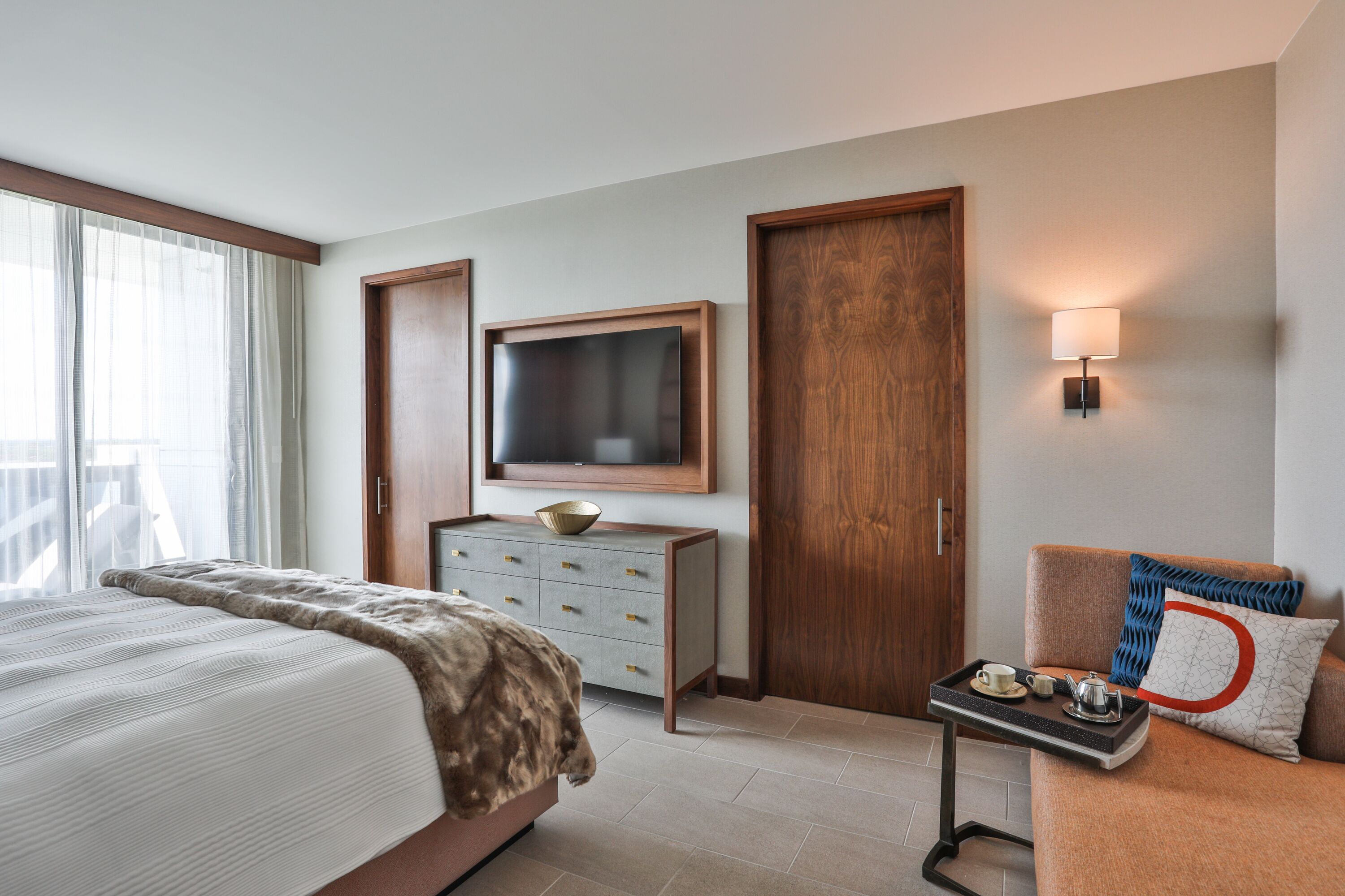 A hotel room with a bed, a dresser, a television, a bedside table with a tray, and a sofa with a pillow.