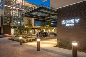 A modern hotel entrance with a glass facade, a visible parking area, and a sign reading "DREY HOTEL".