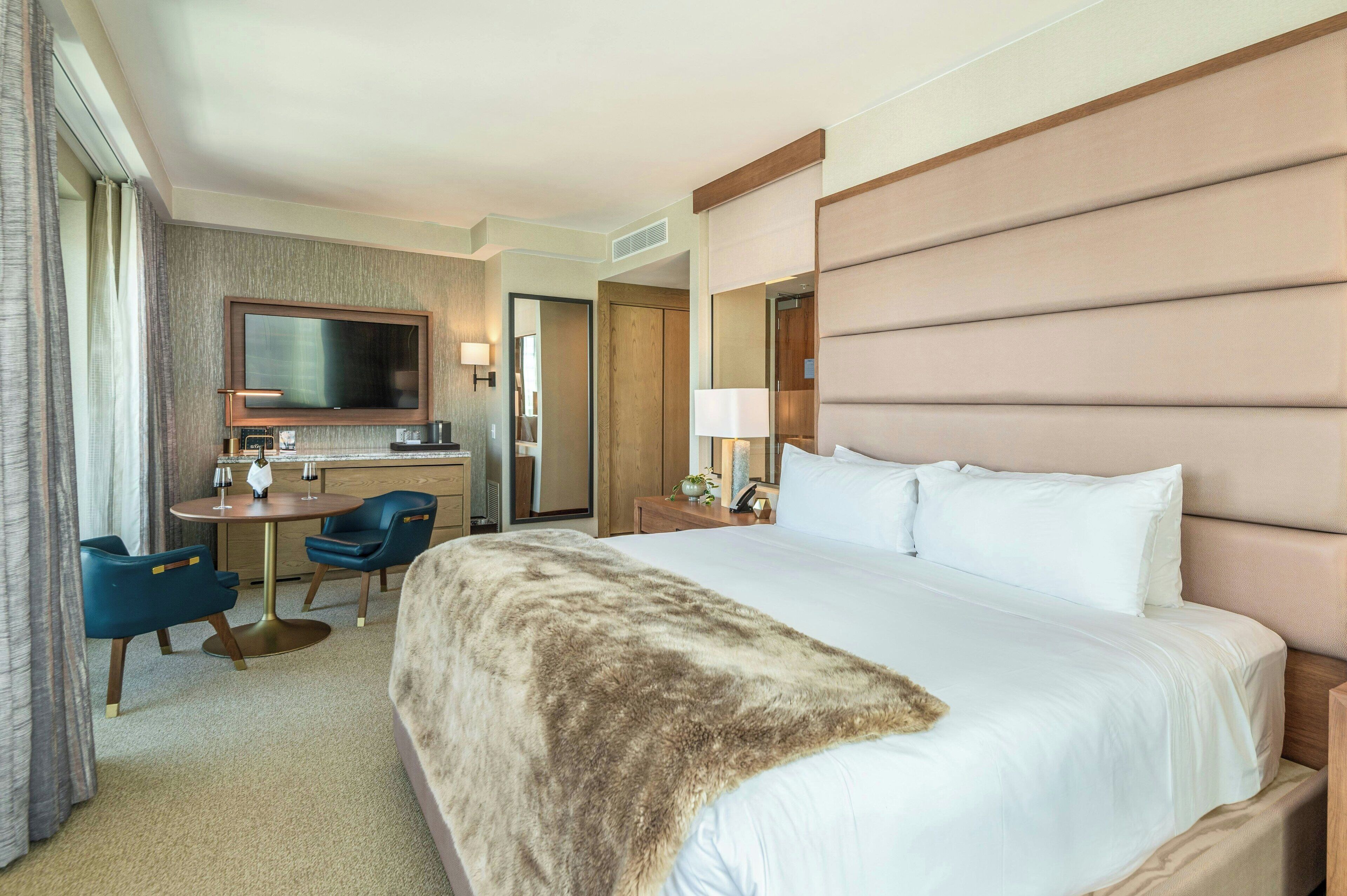 A hotel room with a large bed, a desk with a flat-screen TV, two chairs, a small table, and a mirror.