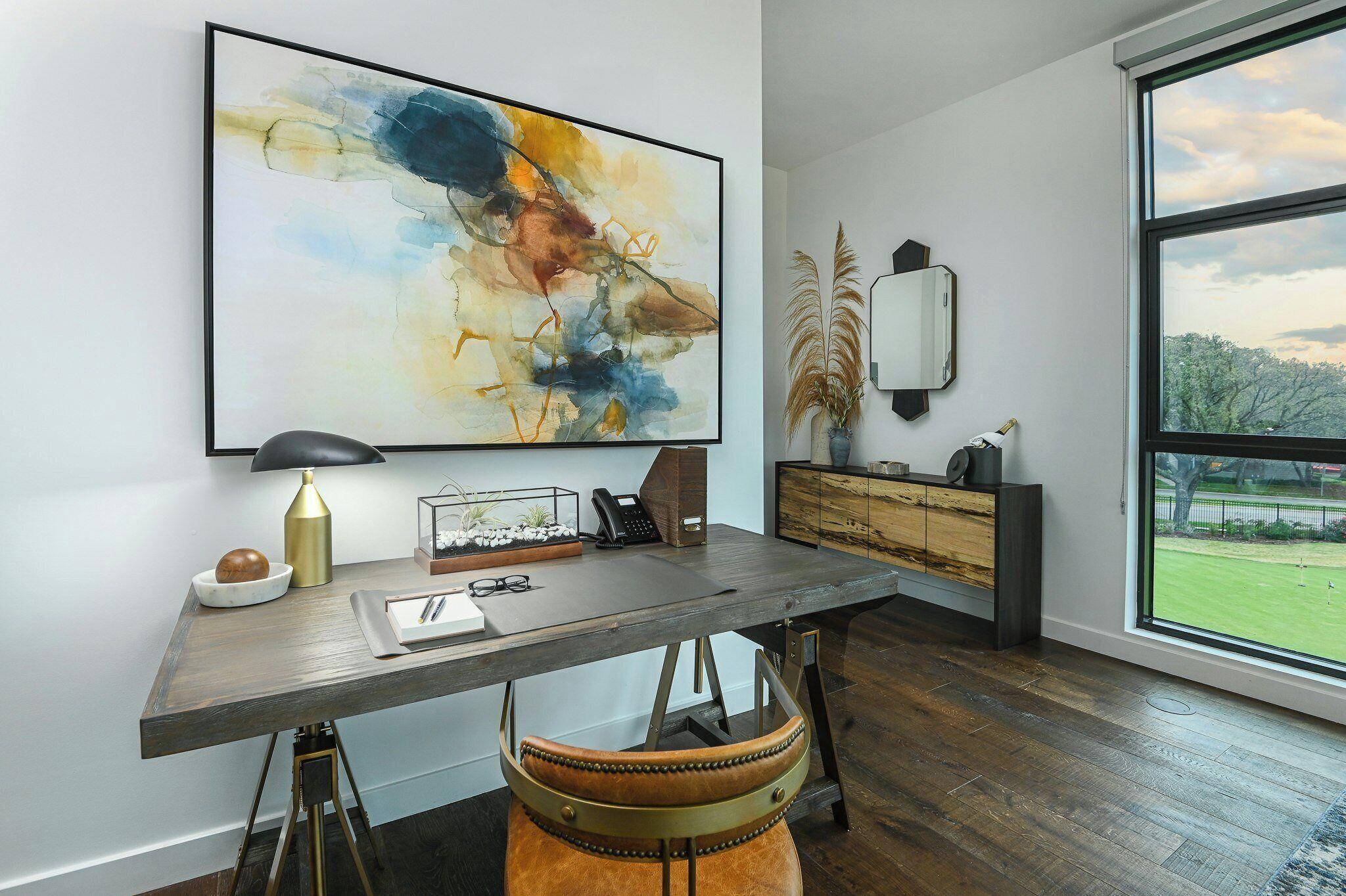 A modern office space with a large abstract painting, a desk with a lamp, a telephone, and a view of a park through a large window.