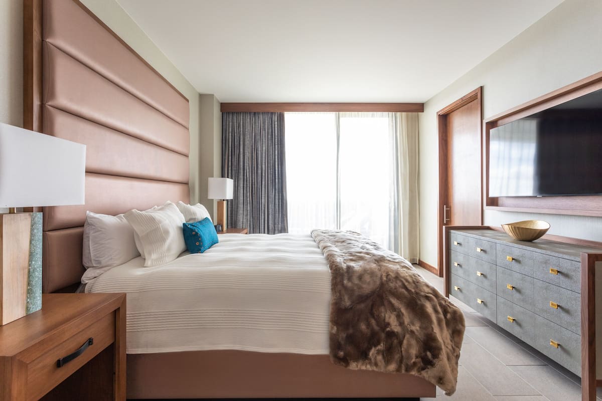 A hotel room with a large bed, a wooden nightstand, a dresser with drawers, a television, and a window with curtains.