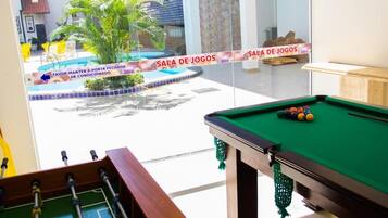 A pool table with a green felt surface and a foosball table in a room with a glass wall.