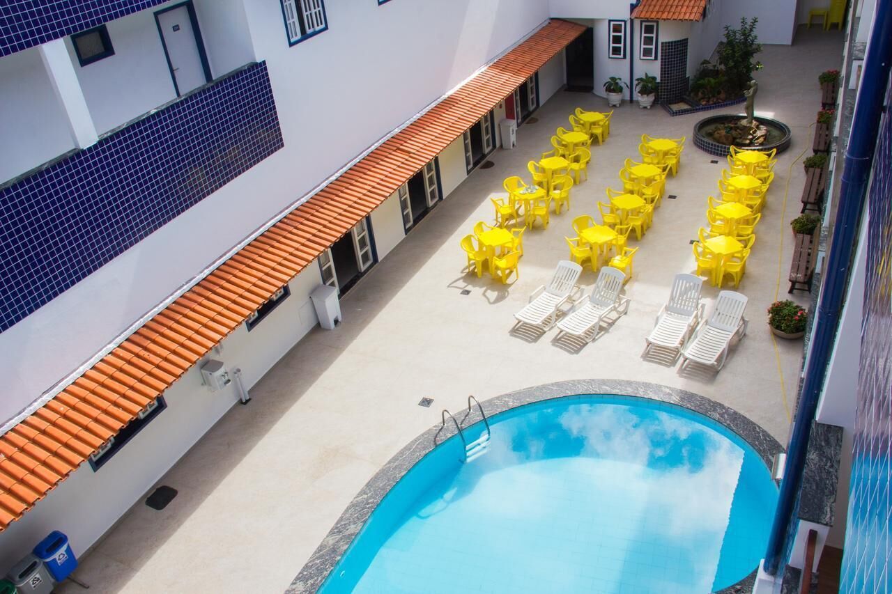 A rooftop pool area with yellow chairs and sun loungers, surrounded by a white building with blue-tiled walls and orange roof tiles.