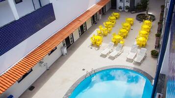 A rooftop pool area with yellow chairs and sun loungers, surrounded by a white building with blue-tiled walls and orange roof tiles.