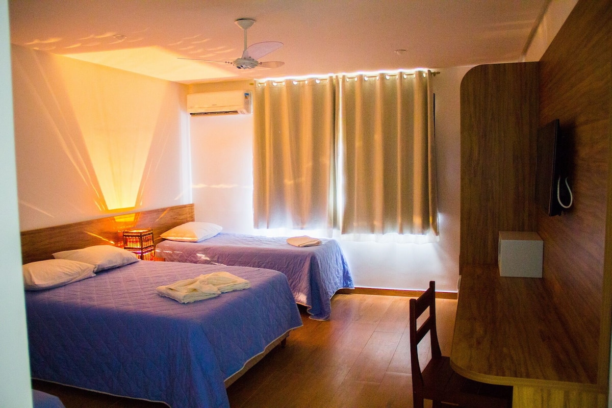 Une chambre d’hôtel avec deux lits, un ventilateur de plafond, un appareil de climatisation, une lampe de chevet, un téléviseur fixé au mur et une fenêtre avec des rideaux.